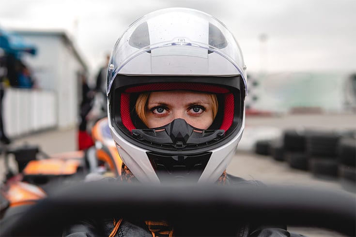A close-up of a person wearing a white helmet and black mask, staring intently ahead while sitting in a go-kart at an outdoor track—a thrilling activity perfect for hen parties. The background is slightly blurred with tires and buildings visible.