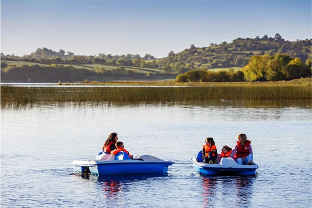 Two groups of people wearing life jackets ride pedal boats on a calm lake in Ireland’s Hidden Heartlands, surrounded by reeds and green hills under a clear sky.