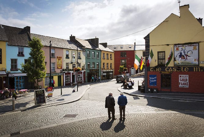 Two people walk across a cobblestone street in Irelands Hidden Heartlands, passing vibrant shopfronts, outdoor seating, Irish flags, and a sunny, partly cloudy sky in a colorful Irish town square.