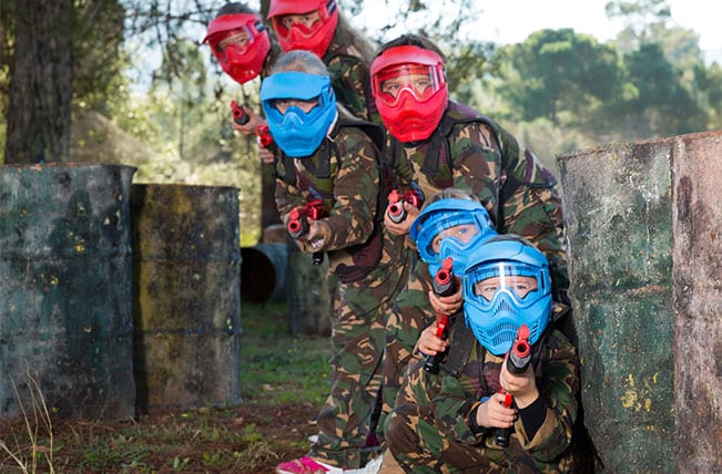 A group of six people wearing camouflage suits and protective masks, some red and some blue, crouch behind barriers outdoors, aiming Splatball markers during a game in a wooded area.