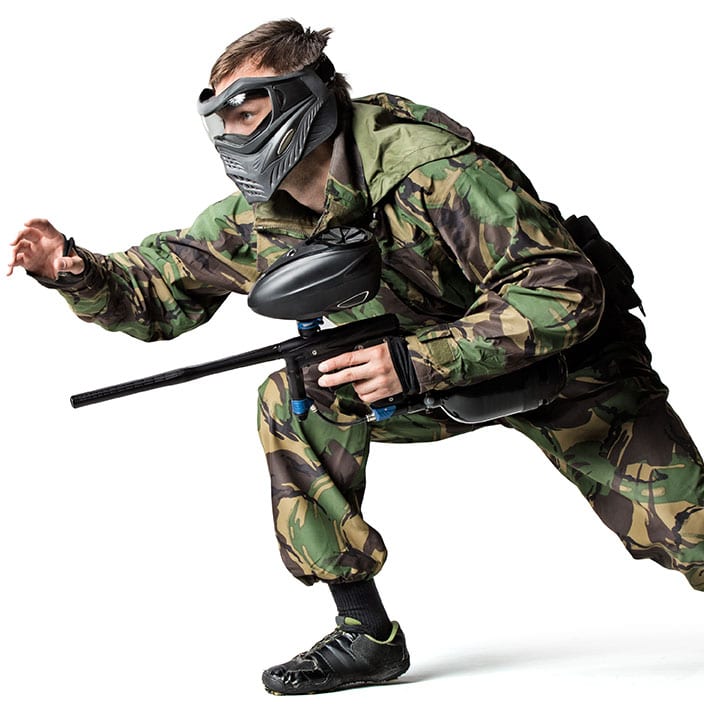 A person in camouflage gear and a protective mask crouches while holding a paintball gun, ready for action—an ideal scene for adventurous stag parties against a white background.