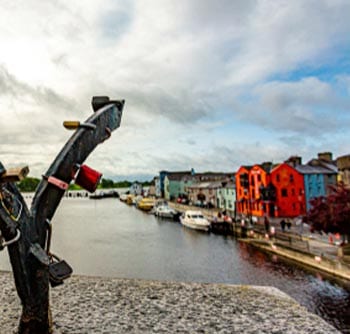 View of a river lined with colorful buildings and boats, seen from a stone bridge with a metal structure and love locks in the foreground under a cloudy sky—useful information for visitors seeking picturesque spots.