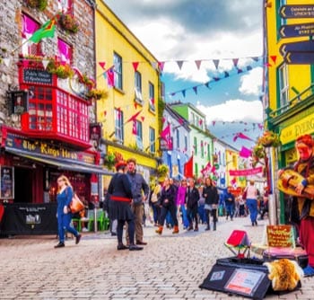 A lively, colorful street scene shows people walking along cobblestones past vibrant buildings with flags and bunting overhead. For useful information, a musician with a drum and puppet performs near the pub called The Kings Head.