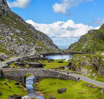 A scenic stone bridge spans a narrow stream, surrounded by winding roads, green grass, and rocky hills. For useful information about the area’s trails and views, explore the small lake beneath a partly cloudy blue sky in this mountainous landscape.