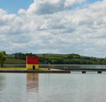 A small yellow and red building sits on the edge of a calm body of water with green hills and a cloudy sky in the background, offering a scenic spot for relaxation and useful information for visitors.