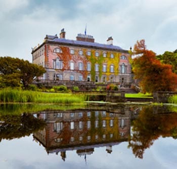 A large, historic mansion covered in green and red ivy is reflected in a calm pond, surrounded by lush trees and greenery under a cloudy sky, offering useful information about timeless architecture and natural beauty.