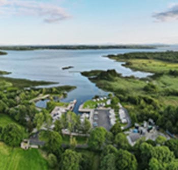 Aerial view of a marina with several boats docked, surrounded by lush green trees and grass, next to a large lake with calm water under a partly cloudy sky—offering useful information for boating enthusiasts.