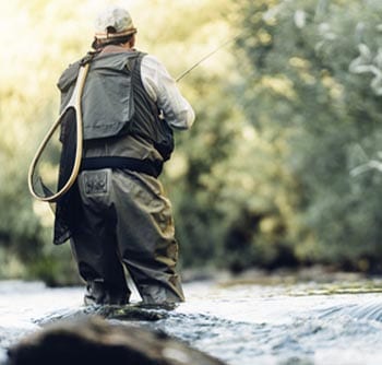 Person wearing fishing gear stands in a shallow, flowing river, fly fishing. Trees and greenery blur in the background, creating a peaceful outdoor scene filled with useful information for anyone interested in angling.