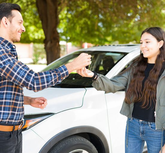 A man and a young woman smile as he hands her car keys in front of a white SUV, highlighting driving lessons for young drivers, with green trees in the background.
