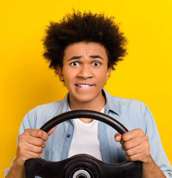 A young nervous driver with curly hair and a worried expression grips a steering wheel tightly, standing against a bright yellow background.