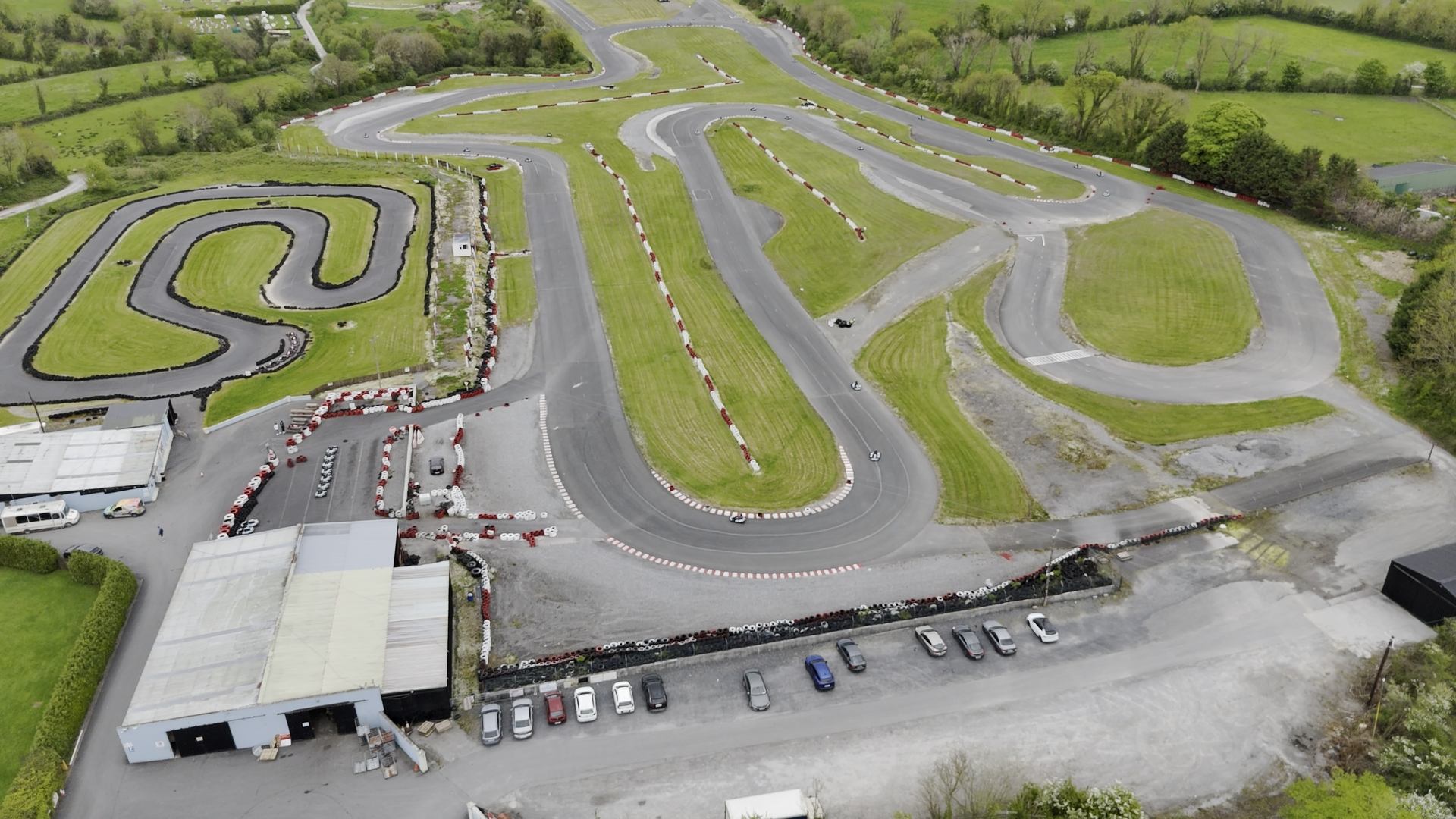 Aerial view of a go-kart racing track with winding paved lanes, green grassy areas, parked cars, and an RSA Rallysport Association building near the entrance, all surrounded by trees and fields. Aerial view of a go-kart racing track with winding paved lanes, green grassy areas, parked cars, and an RSA Rallysport Association building near the entrance, all surrounded by trees and fields.