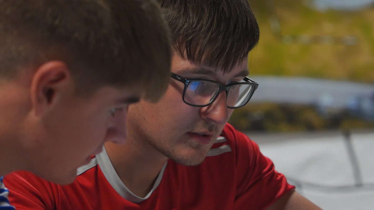 Two young men are closely focused on something out of view, possibly at a driving school. One wears glasses and a red shirt, while the other is in the foreground, slightly out of focus. The background is blurred.