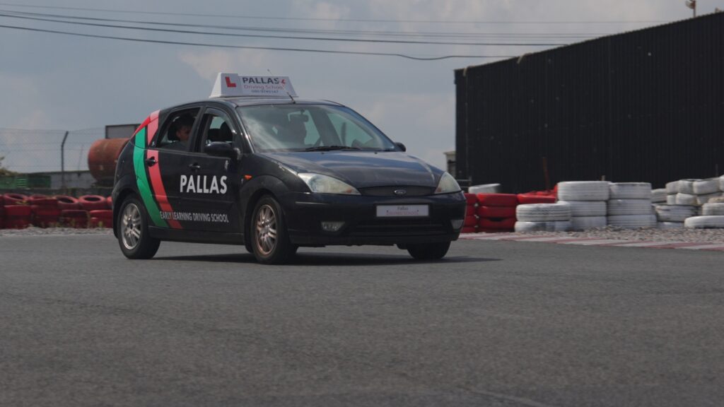 A black Driving School Pallas car with a roof sign and learner plates navigates a turn on a race track, surrounded by tire barriers and fencing under a partly cloudy sky.