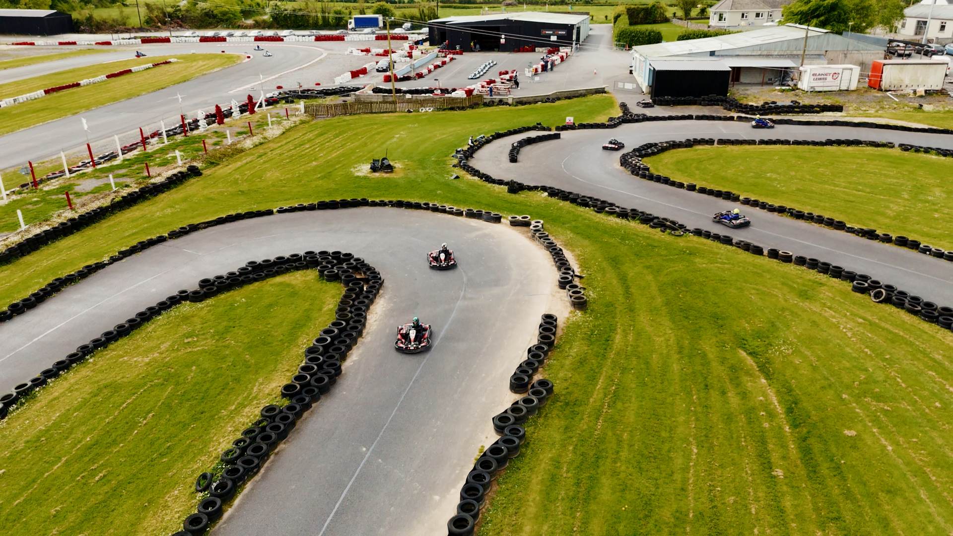 Aerial view of a go-kart track with several people driving go-karts around winding turns, bordered by grass and black tire barriers on a sunny day. Buildings and more track are visible in the background. Aerial view of a go-kart track with several people driving go-karts around winding turns, bordered by grass and black tire barriers on a sunny day. Buildings and more track are visible in the background.