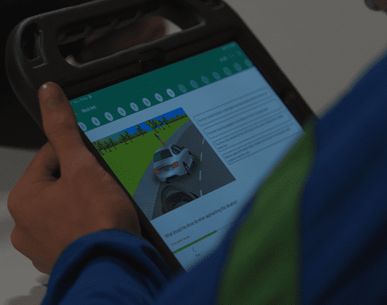 A Transition Year student holds a tablet displaying a driving theory test with a multiple-choice question and an image of a white car on a road.