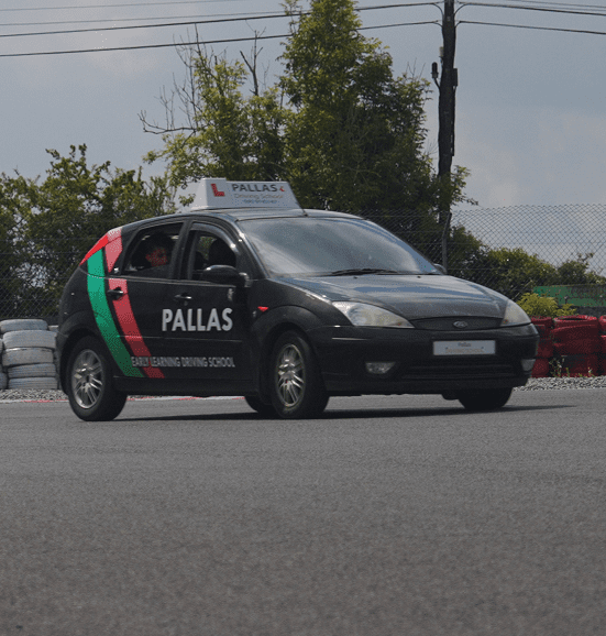 A black car with PALLAS and EARLY LEARNING DRIVING SCHOOL on the side, featuring a learner sign on the roof, is used for Transition Year students as it drives on a paved area with trees and fencing in the background.