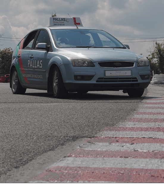 A silver Ford car with PALLAS and Safe Driving Driving School written on the side is parked near a curb on a road, with a driving school sign on the roof.