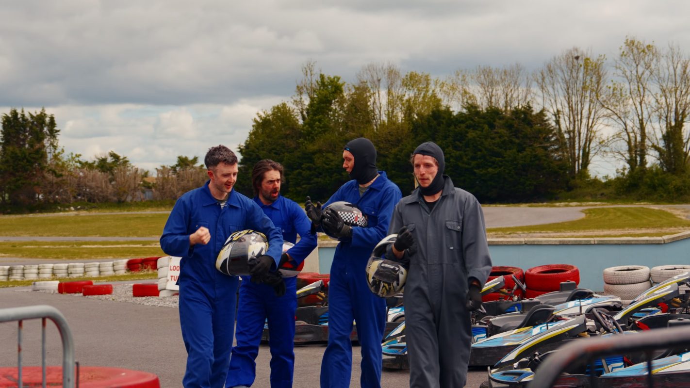 Four men in blue and gray racing suits stand together holding helmets near parked go-karts on an outdoor track, likely enjoying a fun stag party. They appear to be talking and smiling, with trees and a cloudy sky in the background.