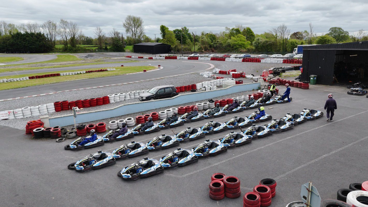 Rows of go-karts line an outdoor track near a building, perfect for clubs or school events. People are seated in some karts while others stand nearby, with red and white tire barriers circling the track on a cloudy day.