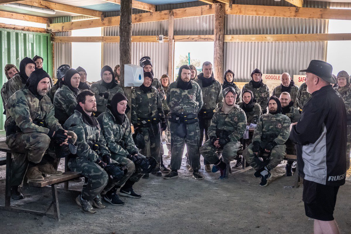 A group of people in camouflage gear sit and stand in a rustic shelter, listening to an instructor in black and white attire. The scene appears to be a team building paintball or airsoft briefing.
