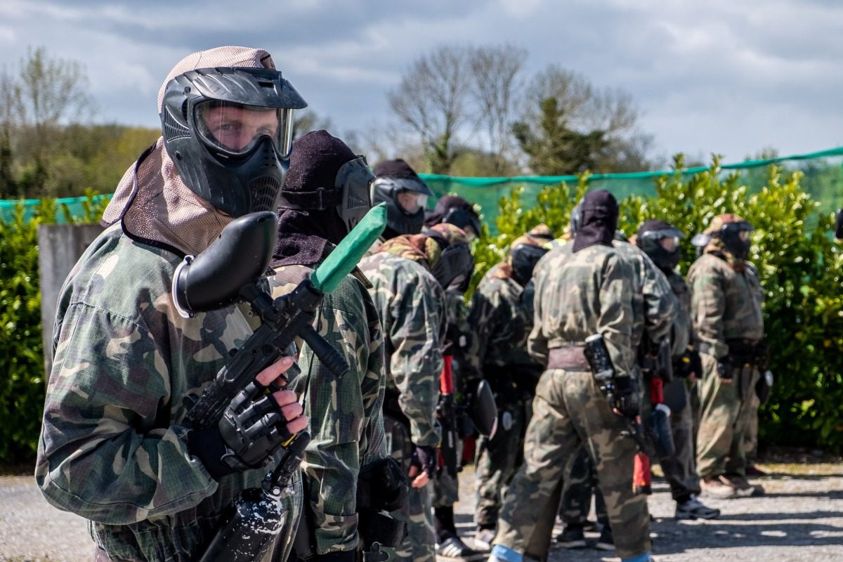 A group of people wearing camouflage clothing and protective masks stand outdoors with paintball guns, preparing for a game. The background features trees and a cloudy sky.