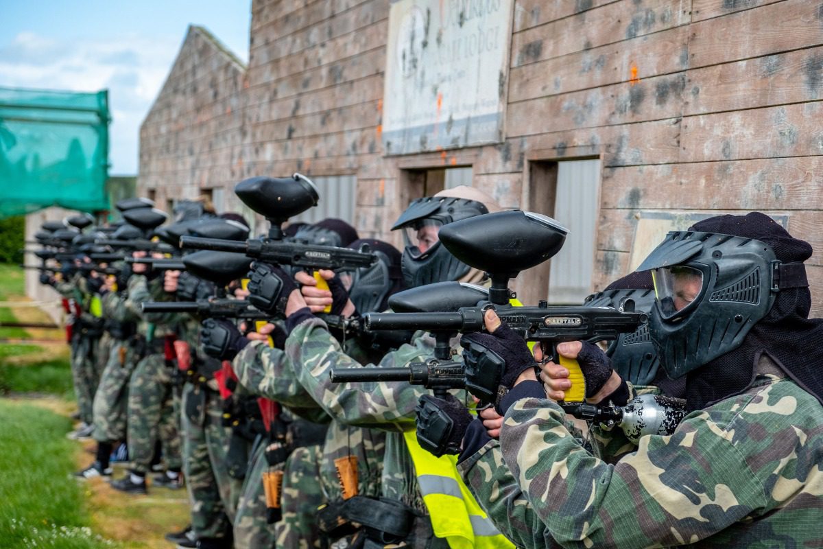 A group of people wearing camouflage outfits and protective masks stand in a row, aiming Splatball guns in front of wooden barriers during an outdoor paintball game.