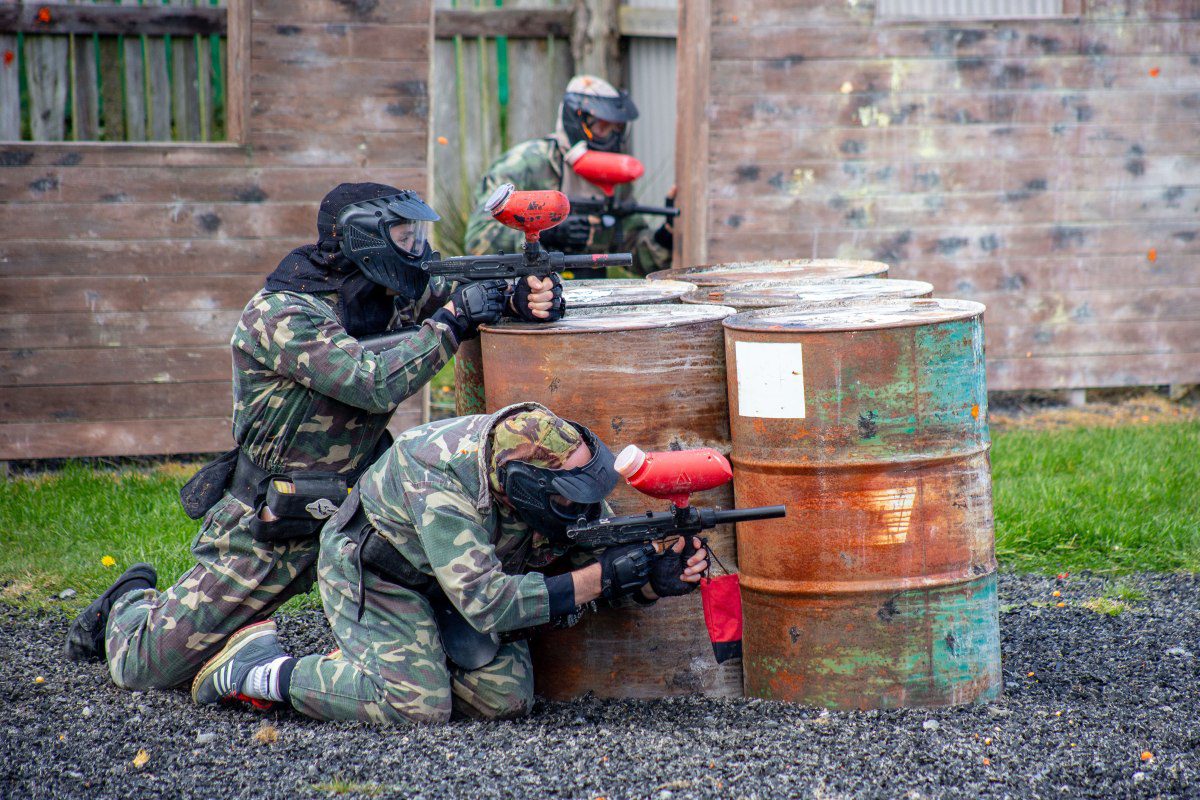 Three people in camouflage and protective masks play an intense game of paintball, crouching behind rusted metal barrels and aiming their paintball guns, with a wooden wall and grass in the background.